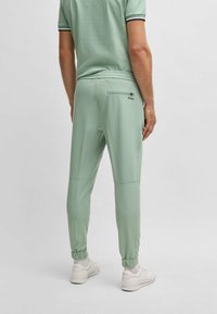 Lichtgroene joggers met een gladde textuur en elastische boorden. Voorzien van een achterzak met rits en een merklogo op de achterkant. Gemaakt met witte sneakers.