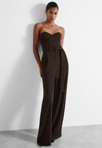 Helfri brun jumpsuit med en åtsittande bodice, vida ben och ett knytbälte i midjan. Slät tyg och strukturerad design.