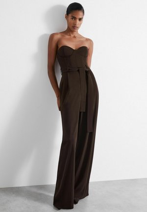 Ruskea olkaimeton jumpsuit, jossa on muotoiltu yläosa, leveät lahkeet ja solmittava vyövyö keskellä. Sujuva kangas ja jäntevä muotoilu.
