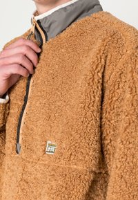 Persona che indossa una giacca in pile color beige con un colletto grigio con zip, tiene il cursore della zip vicino al collo, con un piccolo etichetta di marca visibile.