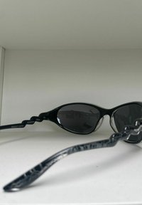 Lunettes de soleil noires avec une monture ovale élégante, des verres sombres et des branches noires texturées arborant un motif subtil. Marquées "KOMONO" et "CAT.3."