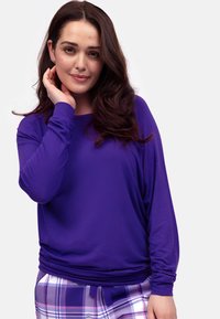 Fioletowa bluzka z długim rękawem, wykonana z lekkiego materiału, o luźnym kroju i marszczonym pasie; zestawiona z spodniami w kratę w delikatnych kolorach.
