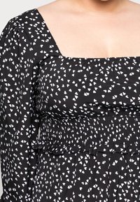 Svart tyg med vita hjärtmotiv, fyrkantig halsringning och åtsittande liv. Smockdetalj i midjan och långa puffärmar.