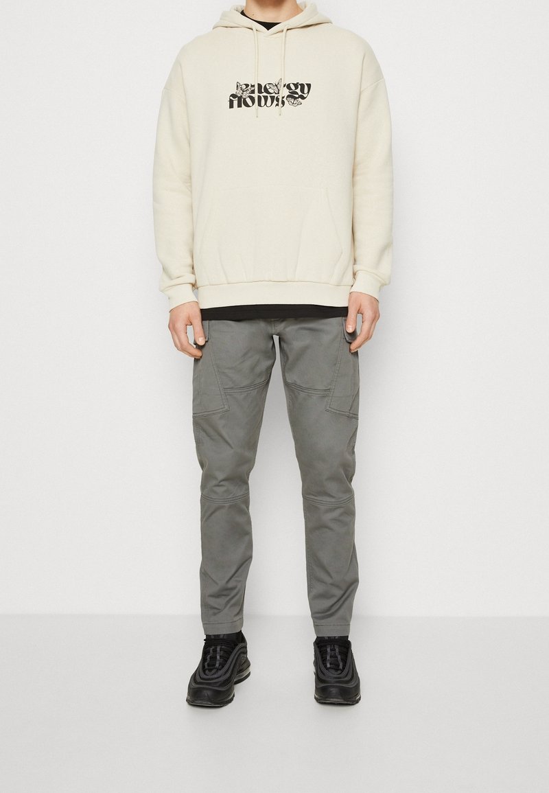 Personne portant un sweat à capuche crème avec un texte noir, un pantalon cargo gris et des baskets noires, debout sur un fond blanc.