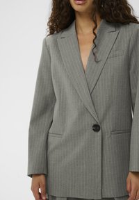 Femeie îmbrăcată într-un sacou gri cu dungi, cu un singur nasture negru, nepurtat, cu mâneci lungi și buzunare frontale vizibile.