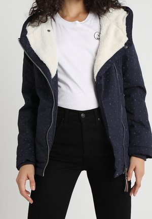 Kobieta w granatowej kurtce z białym futerkiem, białej koszulce i czarnych spodniach z wysokim stanem, stojąca z luźno opuszczonymi rękami.