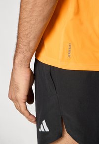 Orangefarbener, kurzärmeliger Sportshirt mit "AEROREADY"-Etikett, kombiniert mit schwarzen Shorts, die ein weißes Adidas-Logo und seitliche Schlitze aufweisen.