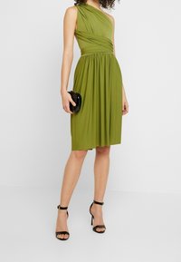 Robe verte mi-longue au genou avec un design asymétrique à une épaule, corsage froncé, jupe drapée, accompagnée d'une pochette noire et de talons à brides.
