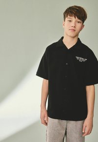 Camisa negra de manga corta abotonada con cuello y texto blanco en el lado izquierdo. Combinada con pantalones claros de color gris con patrón.