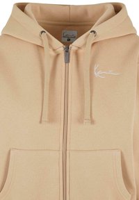Felpa con zip beige chiaro, dotata di cordini, con tasca frontale e logo ricamato. Tessuto morbido con una texture liscia.