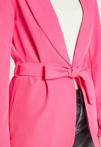 Blazer rose avec une taille ceinturée, fabriqué dans un tissu lisse. Il présente de larges revers et une coupe décontractée, associé à un haut léger en dessous.
