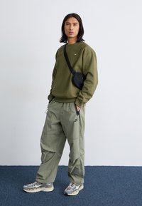 Oseba z dolgimi lasmi stoji samozavestno, oblečena v olivno zeleno majico in cargo hlače, obuta v superge in z majhnim črnim nahrbtnikom.