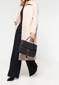 Svart läderhandväska med en strukturerad design, fransdetalj och handtag ovanpå, i kombination med en beige kappa och mörka byxor.