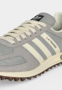 Baskets Adidas L.A. Trainer grises avec des bandes blanches, des lacets beige, des panneaux en mesh et en suède, et une semelle marron texturée.