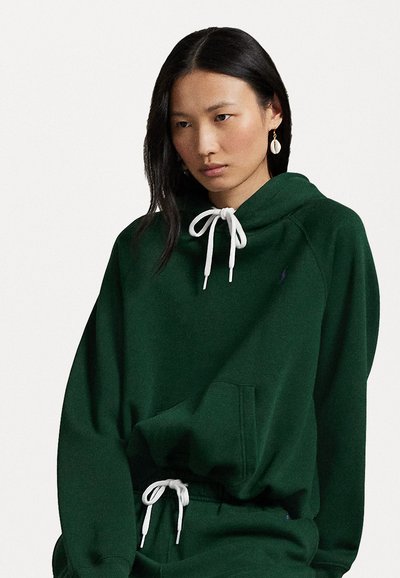 Groene capuchontrui van zacht materiaal, met een voorzak, witte trekkoorden en een klein geborduurd logo op de borst.