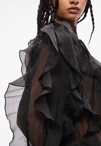 Blusa nera trasparente con strati a ruches lungo le maniche e il corpo, con una texture leggera e fluida e un design con collo alto.