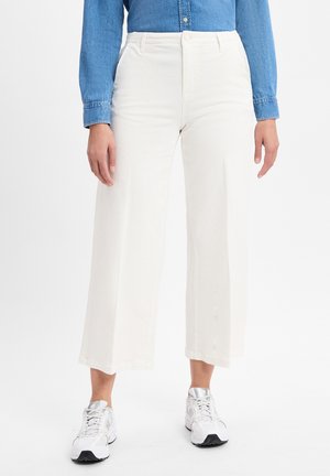 Personne portant un pantalon blanc large coupé, une chemise en jean bleue et des baskets blanches, se tenant sur un fond blanc.