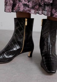 Bottines noires en cuir gaufré crocodile avec un talon fin, dotées d'un détail de fermeture éclair dorée et d'un design à bout pointu.