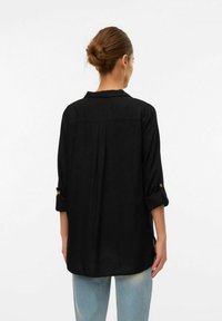Schwarze Button-Down-Bluse mit hochgerollten Ärmeln, Kragen und einem Rückenfalte-Detail. Der Stoff wirkt glatt; die Bluse hat eine lockere Passform und ein hüftlanges Design.