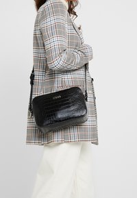 Žena v kostkovaném saku a bílých kalhotách nese černou crossbody tašku s krokodýlí strukturou a viditelným logem značky.