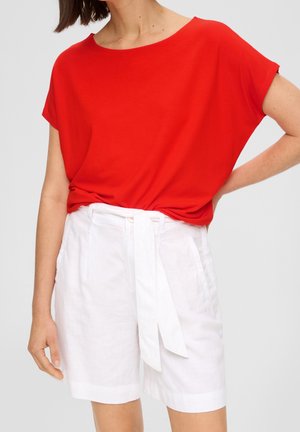 Vrouw draagt een felrode korte mouwen blouse, gestopt in witte shorts met hoge taille en riem, met zakken, staat met één hand op de heup.
