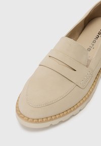 Chaussure à enfiler en suede beige avec un capuchon de chaussure cousu, des accents de fente doubles et une semelle en caoutchouc texturée. Design minimaliste sans logos visibles.