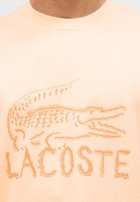 Κοντινό πλάνο ενός πουκάμισου σε χρώμα ροδακινί με κεντημένο πορτοκαλί κροκοδείλιο και τη λέξη "LACOSTE" κάτω από το σχέδιο.