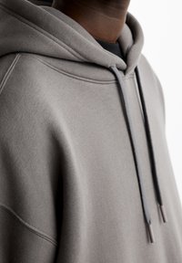 Šedá mikina s měkkou texturou, vybavená kapucí na šňůrku a detailním prošitím na bočních švech. Materiál je hladký a má uvolněný střih.