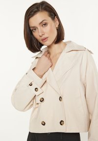 Lichtbeige cropped jasje met een dubbelzijdig ontwerp, grote bruine knopen, brede kraag en zijzakken. Glad textuur van de stof.