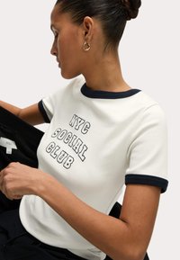 Witte katoenen t-shirt met een marineblauwe geribde kraag en mouwen. Voorzien van geborduurde tekst "NYC SOCIAL CLUB". Model houdt een zwart item vast.
