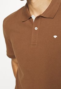 Brązowa koszulka polo z teksturowanej tkaniny, z dwoma guzikami z przodu, krótkimi rękawami i małym białym logo na piersi.