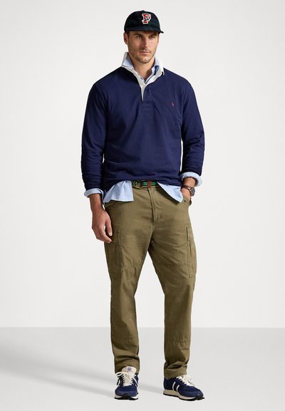 Pulover bleumarin cu guler peste o cămașă bleu deschis, pantaloni cargo oliv, adidași negri și o șapcă închisă cu un logo.