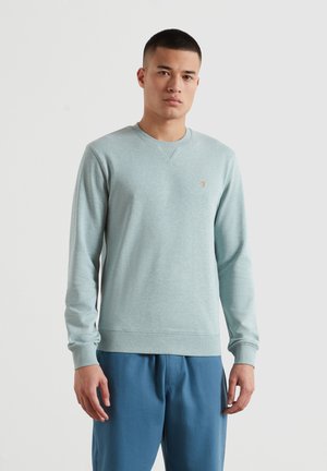 Mężczyzna o krótkich włosach stojący, ubrany w jasnoniebieską bluzę z okrągłym dekoltem i niebieskie spodnie na jednolitym białym tle.