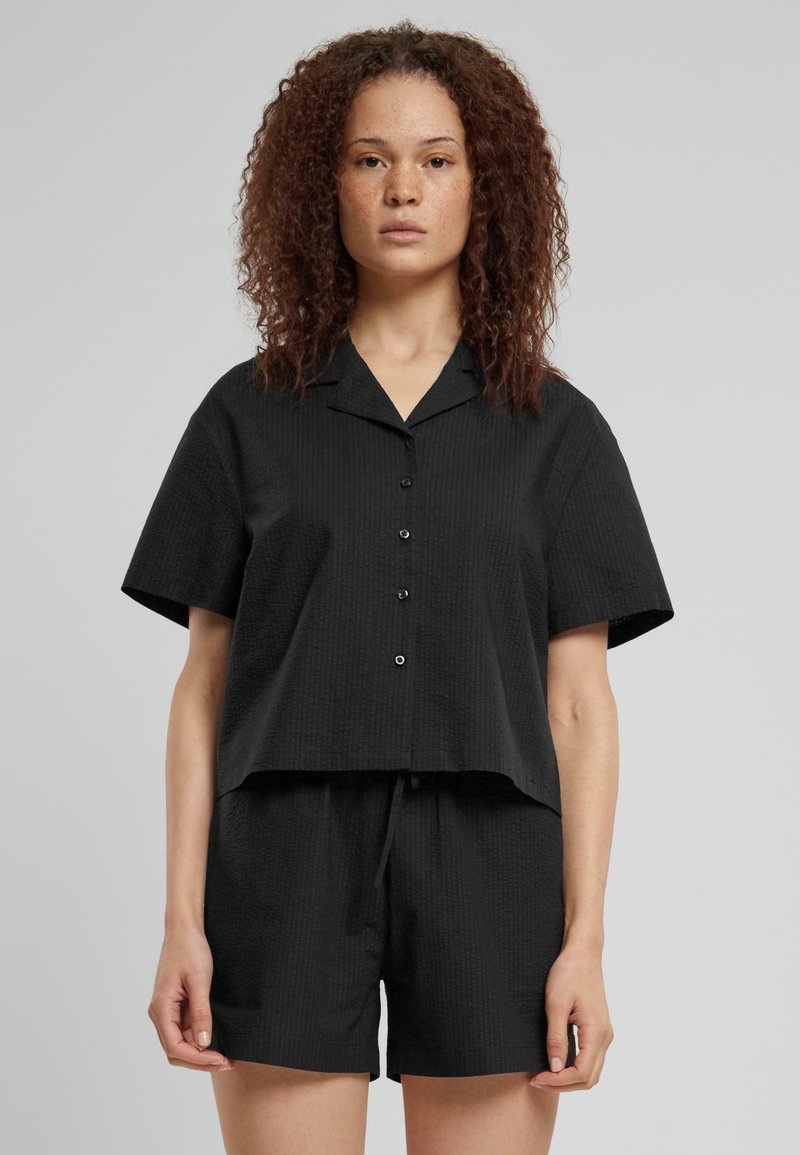 Maglietta nera a maniche corte con colletto e chiusura a bottoni, abbinata a pantaloni corti neri coordinati. Entrambi presentano una finitura in tessuto con texture.