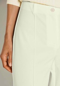 Close-up van een hand die rust naast hooggetailleerde, crèmekleurige broek met knoopdetails en een tailleband.