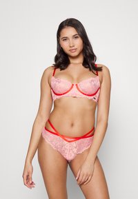 Conjunto de lingerie em renda na cor rosa claro com realces em vermelho. O sutiã apresenta aro e renda transparente, enquanto as calcinhas têm tiras entrelaçadas.