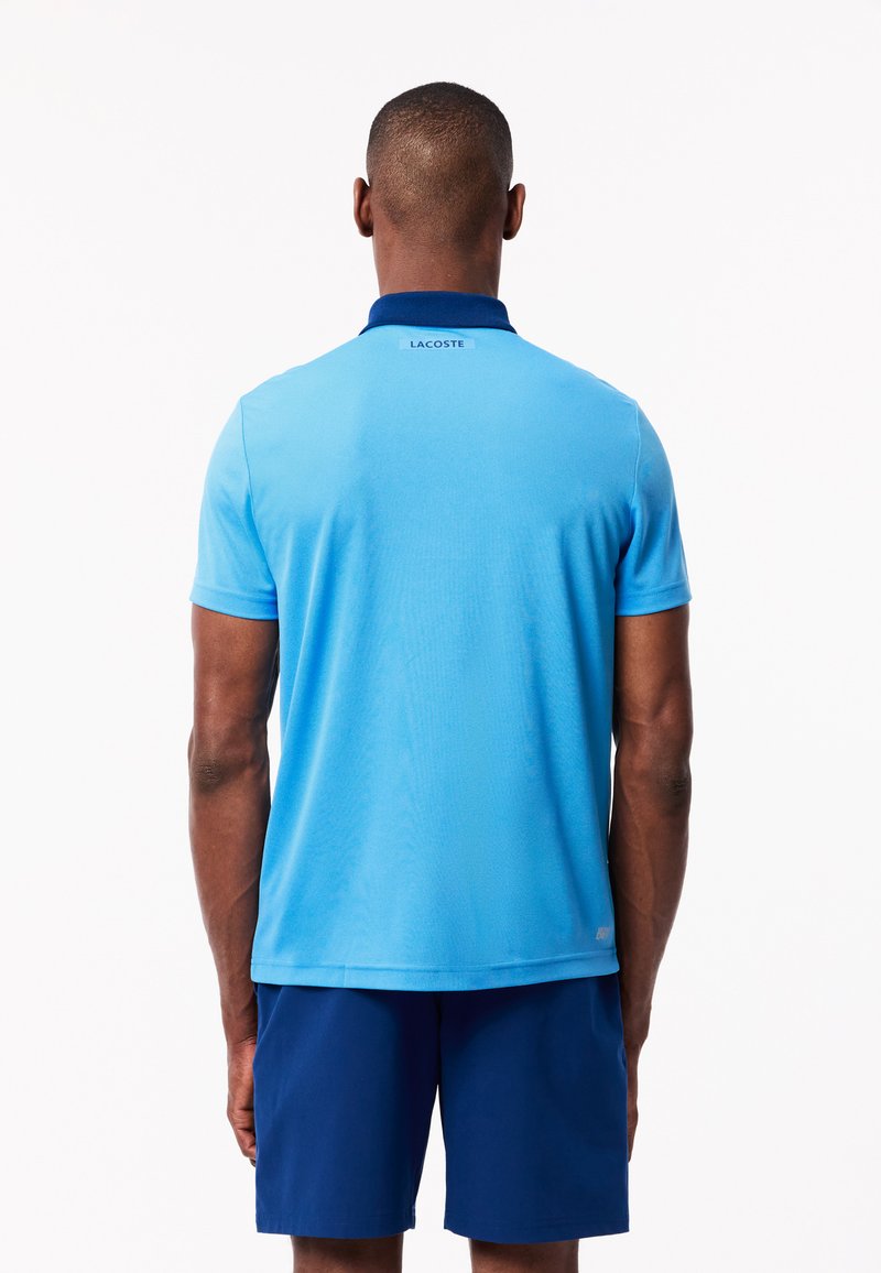 Polo léger bleu clair avec un col marine, manches courtes et une texture subtile, arborant le logo "LACOSTE" au dos.