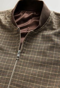 Brown check-patterned bomber jacket with a silver zipper and ribbed collar, featuring a smooth inner lining in a darker brown shade.