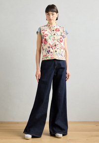 Blommönstrad väst med fickor, beige bakgrund och knappstängning, ihop med vida mörk denimbyxor och vita sneakers.