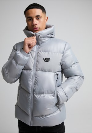 Joven vestido con chaqueta acolchada gris claro con cuello alto, sosteniendo la cremallera cerca del cuello, de pie frente a un fondo liso.
