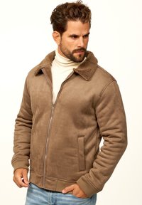 Braune Wildleder-Bomberjacke mit einem strukturierten Futterkragen aus Kunstschurwolle, Reißverschluss, Seitentaschen und gerippten Bündchen. Getragen über einem beigen Rollkragenpullover.