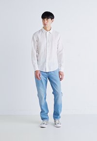 Camicia bianca in lino a maniche lunghe con chiusura a bottoni, abbinata a jeans di denim azzurri chiari. Sneakers casual in bianco e nero completano l'outfit.