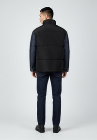 Czarna pikowana kamizelka z wysokim kołnierzem, jeansowymi rękawami i kontrastującymi guzikami, w zestawieniu z ciemnoniebieskimi spodniami i czarnymi butami.