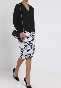 Blouse à manches longues noire avec garniture en satin, associée à une jupe crayon mi-longue à fleurs en noir et blanc. Accessoirisée d'une pochette noire.