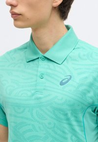 Jeune homme portant un polo turquoise à motifs avec un petit logo sur la poitrine gauche, vu du cou jusqu'au milieu du torse.