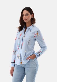 Light blue button-up shirt with colorful fashion illustrations. Features a collar, long sleeves, and striped cuffs. Made from smooth fabric.