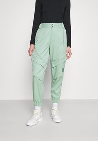 Lichtgroene joggerbroek met een losse pasvorm, voorzien van elastische boorden, cargozakken en twee knopen aan de voorkant. Het materiaal lijkt glad en veelzijdig.