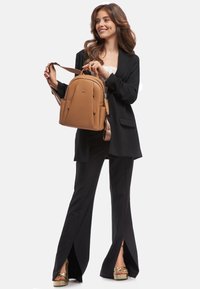 Zaino in pelle marrone con finitura testurizzata, doppie tasche laterali, spallacci regolabili e manico superiore. Indossato con un blazer nero e pantaloni con spacco.