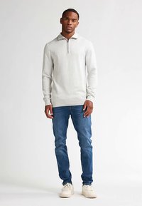 Jasnoszary sweter z długim rękawem i kołnierzem na pół zamek, zestawiony z niebieskimi dżinsami i jasnymi trampkami. Gładka faktura i dopasowany krój.