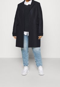 Henkilö pukeutuneena mustaan villatakkiin mustan neuleen ja valkoisen paidan päällä, vaaleansinisiin farkkuihin ja valkoisiin lenkkareihin, seisoo harmaalla lattialla.